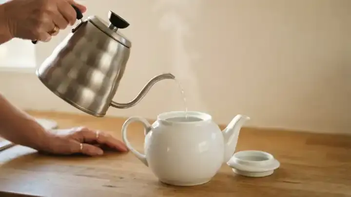 A kettle pouring steaming water into a white porcelain teapot against a soft plain cream wall, slow zoom